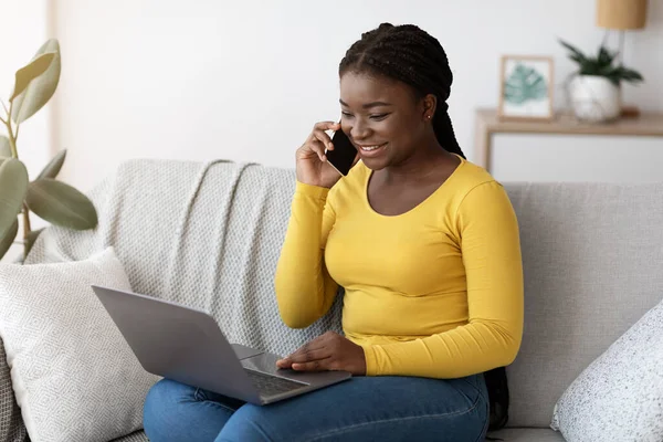 Sorridente donna nera che parla sul cellulare e utilizzando il computer portatile a casa — Foto Stock
