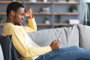 Dijital tabletle kanepede oturan düşünceli siyah adam.