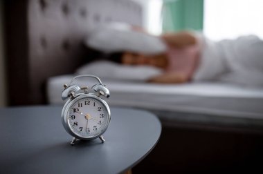 Komodindeki çalar saat ve uyanmak istemeyen genç kadının bulanık görüntüsü. Yatakta yastıkla kulaklarını örtüyor.