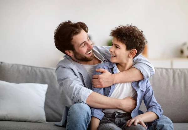 Pojęcie dwóch pokoleń. Szczęśliwy ojciec obejmujący syna, patrzący na siebie i uśmiechający się, siedzący na kanapie w domu — Zdjęcie stockowe