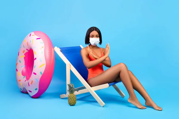 Proibição de viajar pelo Coronavirus. Mulher negra em maiô e máscara facial mostrando gesto PARAR enquanto sentada na cadeira lounge — Fotografia de Stock