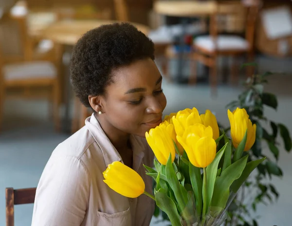 Portrait of beautiful black lady smelling bouquet of yellow tulips at coffee shop