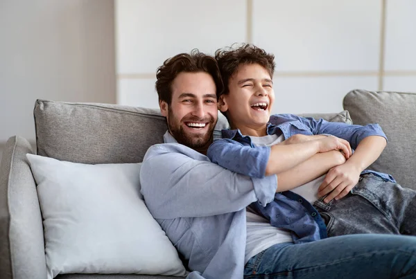 Concept de paternité. Père joyeux embrassant fils, chatouillant et riant tout en se reposant sur le canapé à la maison dans le salon — Photo