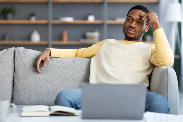 Pensativo hombre de negocios negro sentado en el sofá delante de la computadora portátil —  Fotos de Stock