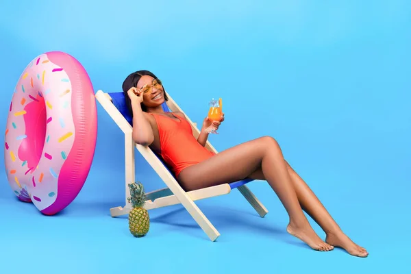 Portrait complet de jeune femme noire se refroidissant dans une chaise longue avec un délicieux cocktail tropical sur fond bleu — Photo