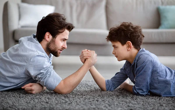 Воспитывать настоящего мужчину. Концентрированные отец и сын армрестлинг и конкурирующие, лежа на полу — стоковое фото