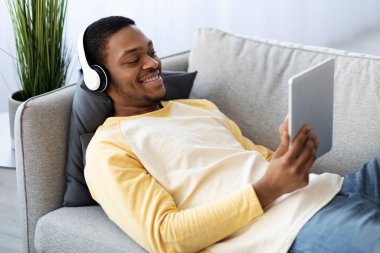 Neşeli siyah adam kanepede uzanıyor, yastıkta film seyrediyor.