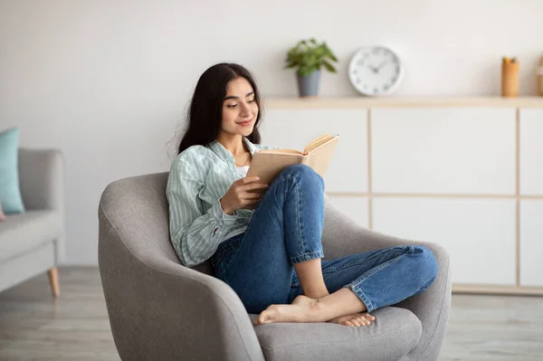 Fique em casa passatempos. Millennial mulher indiana sentado em poltrona aconchegante com livro aberto dentro de casa — Fotografia de Stock