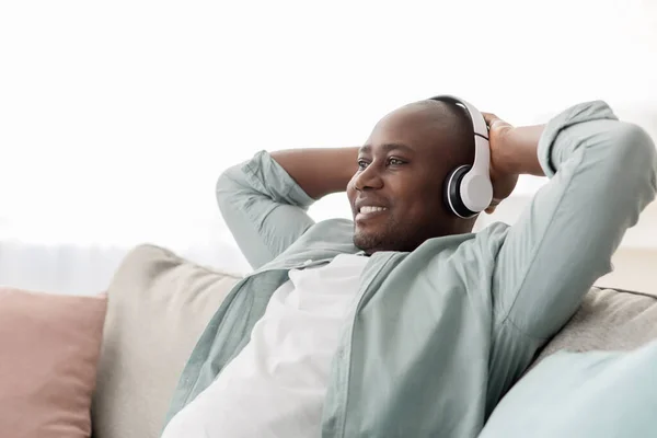 Afrikanischer Mann mittleren Alters in drahtlosen Kopfhörern, der Musik auf dem Sofa im Wohnzimmer hört, Kopierraum — Stockfoto