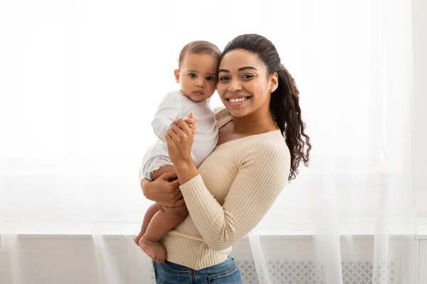 Preto mãe segurando pouco infantil sorrindo para câmera posando dentro — Fotografia de Stock