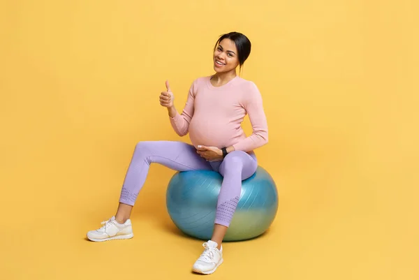 Lycklig gravid svart dam tränar på fitball, tittar på kameran och visar tummen upp, träning på gul bakgrund — Stockfoto