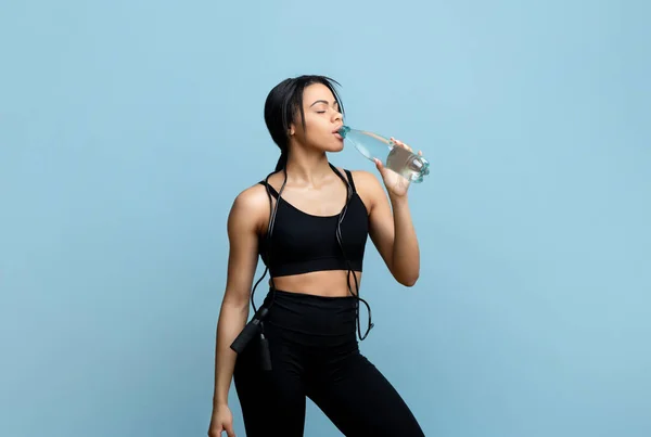 Sport hydratatie concept. Sportieve Afro-Amerikaanse vrouw drinken mineraalwater uit fles op blauwe studio achtergrond — Stockfoto