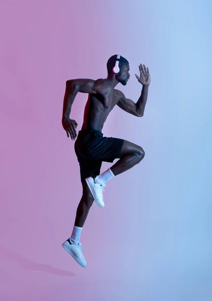 Longitud completa de deportista atlético negro en auriculares corriendo y escuchando música en iluminación de neón, vista lateral —  Fotos de Stock