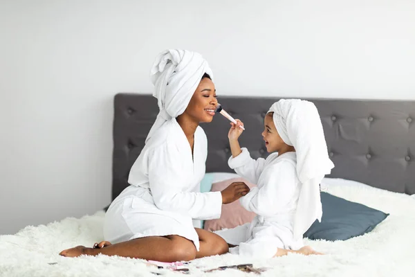 Retrato de comprimento total de mãe e filha afro-americana em roupões de banho e toalhas aplicando maquiagem na cama em casa — Fotografia de Stock
