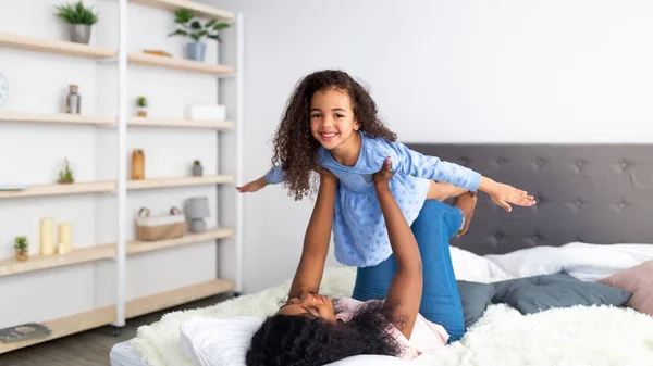 快乐的黑人妈妈和她可爱的女儿在床上玩飞机游戏，全景。家庭团聚时间 — 图库照片