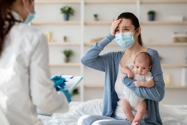 Мать в защитной маске держит новорожденного ребенка, объясняя симптомы болезни врачу — стоковое фото