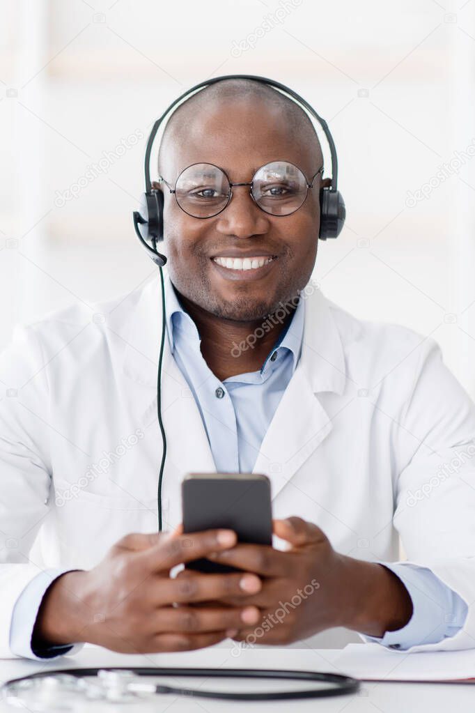 Hot line medical center. Friendly black male doctor in headphones holding smartphone in hand