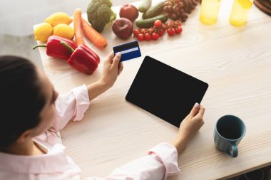 Mutfakta tablet kullanan, elinde kredi kartı olan bir kadın.