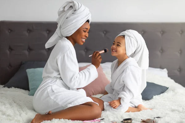 Happy black mom and her cute daughter wearing bath robes and towels, applying makeup on family spa day at home