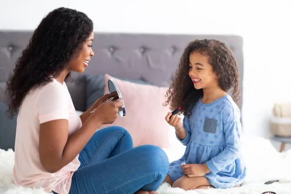 Ημέρα οικογενειακής ομορφιάς. Χαριτωμένο μικρό μαύρο κορίτσι εφαρμογή κραγιόν, ενώ η μαμά της κρατώντας καθρέφτη στο κρεβάτι στο σπίτι — Φωτογραφία Αρχείου