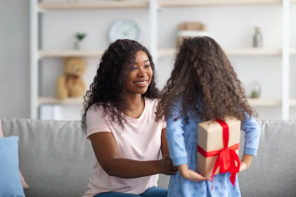 Pequeña niña negra escondida envuelta caja de regalo para las vacaciones a sus espaldas, felicitando a su madre con cumpleaños en casa — Foto de Stock