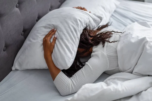 Mujer negra estresada cubriendo la cabeza bajo la almohada acostada en la cama —  Fotos de Stock