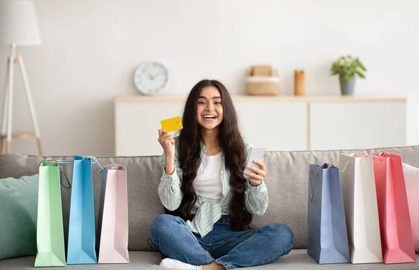 Kredi kartı ve akıllı telefonlu olumlu Hintli kadın evdeki alışveriş torbalarıyla çevrili kanepede oturuyor. — Stok fotoğraf