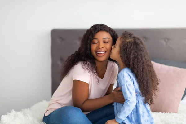 Ένα τρυφερό μαύρο κορίτσι να φιλάει τη μητέρα της στο κρεβάτι στο σπίτι. Οικογενειακοί χρόνοι σύνδεσης — Φωτογραφία Αρχείου