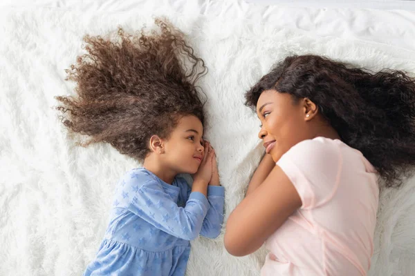 Μικρή μαύρη κοπέλα με τη μαμά της με τα καθημερινά της ρούχα ξαπλωμένη στο κρεβάτι, κοιταζόμενη η μία την άλλη, με θέα στην κορυφή — Φωτογραφία Αρχείου