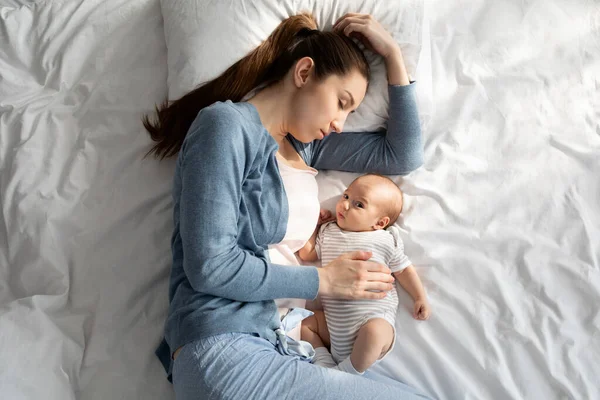 Zmęczona młoda matka padająca w łóżku, gdy jej urocze nowonarodzone dziecko się budzi — Zdjęcie stockowe
