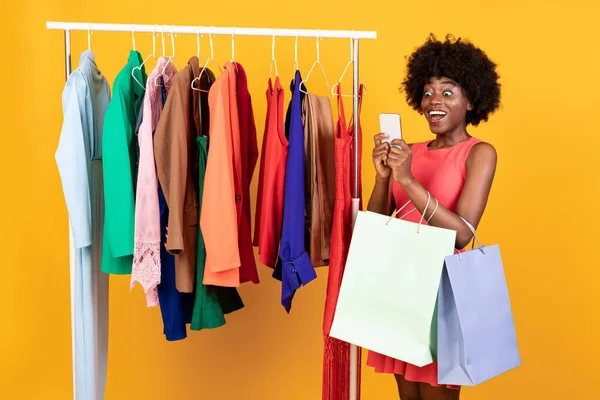 Zwarte dame met behulp van telefoon winkelen in de buurt van Kleding Rail, Gele achtergrond — Stockfoto