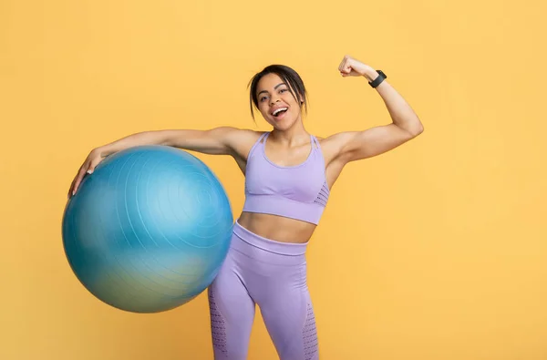Positiv afrikansk amerikansk smal kvinna i sportkläder håller fitness boll och visar biceps på gul bakgrund — Stockfoto