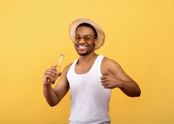 여름 옷을 입고 맥주 병을 들고 노란색 스튜디오 배경에서 엄지손가락 동작을 하는 잘생긴 흑인 남자 — 스톡 사진