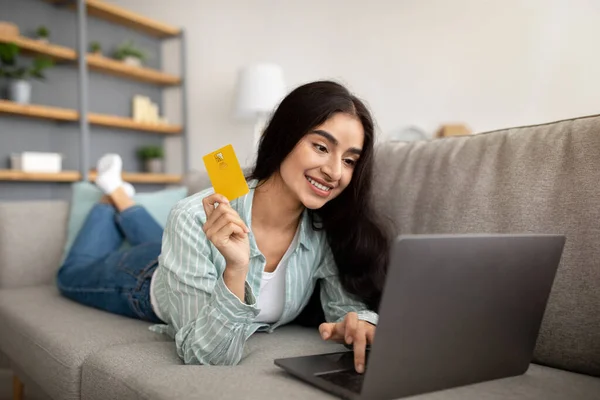Online shopping. Beautiful Indian lady with laptop and credit card lying on sofa at home, buying goods in web store
