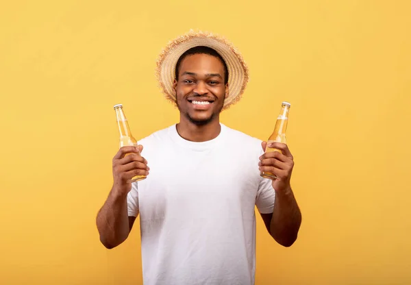 Jovem afro-americano alegre segurando duas garrafas de cerveja e sorrindo para a câmera no fundo do estúdio amarelo — Fotografia de Stock