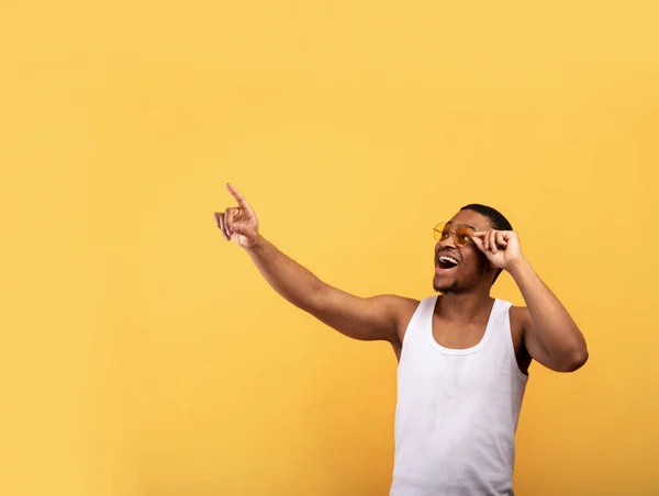 Chico negro excitado con ropa de verano y gafas de sol apuntando al espacio vacío sobre fondo amarillo. Concepto de venta promocional — Foto de Stock