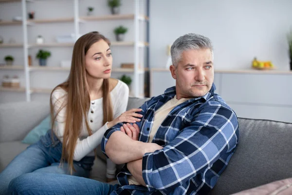 Familienkonfliktkonzept. Verärgerter reifer Mann sitzt auf Couch, seine Frau versucht, nach einem Kampf zu Hause Frieden zu schließen — Stockfoto
