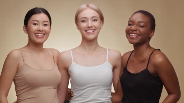 Bellezza femminile diversa. Tre giovani donne felici e diverse che si abbracciano e ridono alla macchina fotografica, sfondo beige — Video Stock