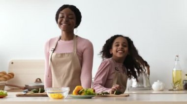 Mutlu Afro-Amerikan anne ve kızı önlük giyip birlikte dans ediyor, mutfakta yemek pişiriyor ve gülüyorlar.