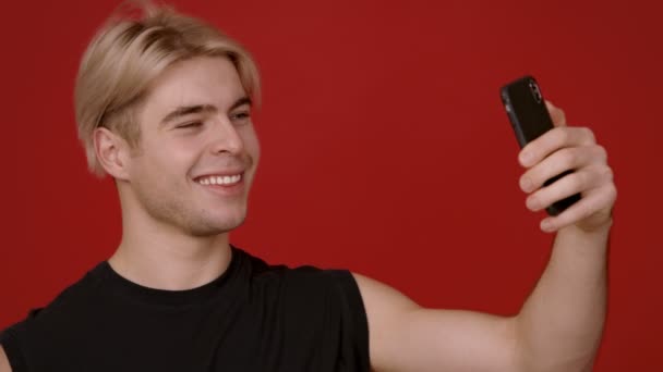 Jovem com cabelo loiro tomando selfie ob smartphone, sorrindo e mostrando v sinal para câmera de celular, fundo vermelho — Vídeo de Stock