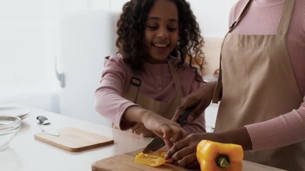 Nettes afrikanisch-amerikanisches Mädchen, das ihr beim Kochen hilft, dabei zusieht, wie Mama Paprika schneidet und Scheiben isst, in der Küche tanzt — Stockvideo