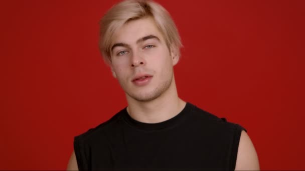Studio portrait d'un mec blond confiant souriant à la caméra et prétendant ses cheveux, fond rouge, ralenti — Video