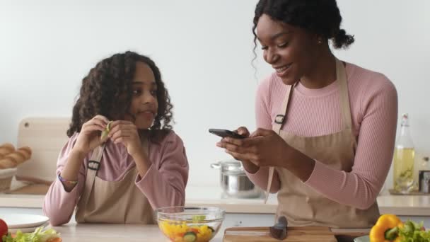 Happy african american mother and daughter making picture of vegetable salad on smartphone for social media at kitchen — Stock Video