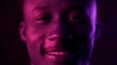 Mor neon ışıklarıyla kameraya gülen mutlu Afro-Amerikalı genç adamın yakın portresi.