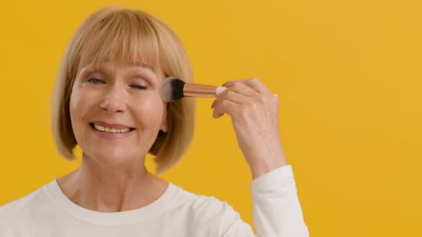 Positieve senior dame het aanbrengen van rouge op de wang met make-up borstel, glimlachen voor de camera, oranje achtergrond met lege ruimte — Stockvideo