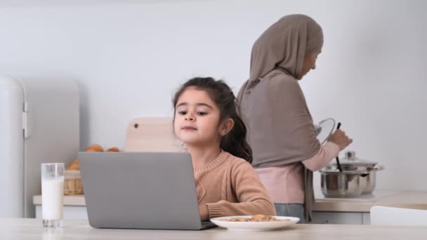 Hija musulmana teniendo clase en línea mientras madre cocina en la cocina — Vídeos de Stock