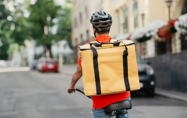 Jovem mensageiro masculino em camiseta laranja, capacete de segurança com mochila térmica amarela, passeios de bicicleta na estrada de rua — Fotografia de Stock