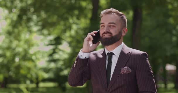 Всегда на связи. Успешный бизнесмен в разговоре по смартфону, стоя в городском парке, свободное пространство — стоковое видео
