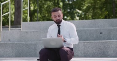 Orta yaşlı iş adamı doğayı seviyor, dizüstü bilgisayarla çalışıyor, parkta merdivenlerde oturuyor ve dans ediyor.