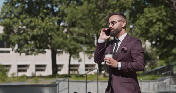 Зайнятий зрілий бізнесмен розмовляє по мобільному телефону, п'є каву під час прогулянки в весняному міському парку — стокове відео